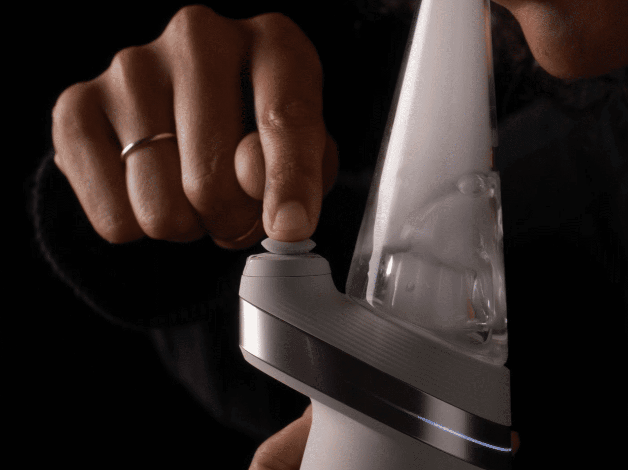 A user pressing the button on a white Puffco electric dab rig to engage the heating element while preparing to inhale vapor.